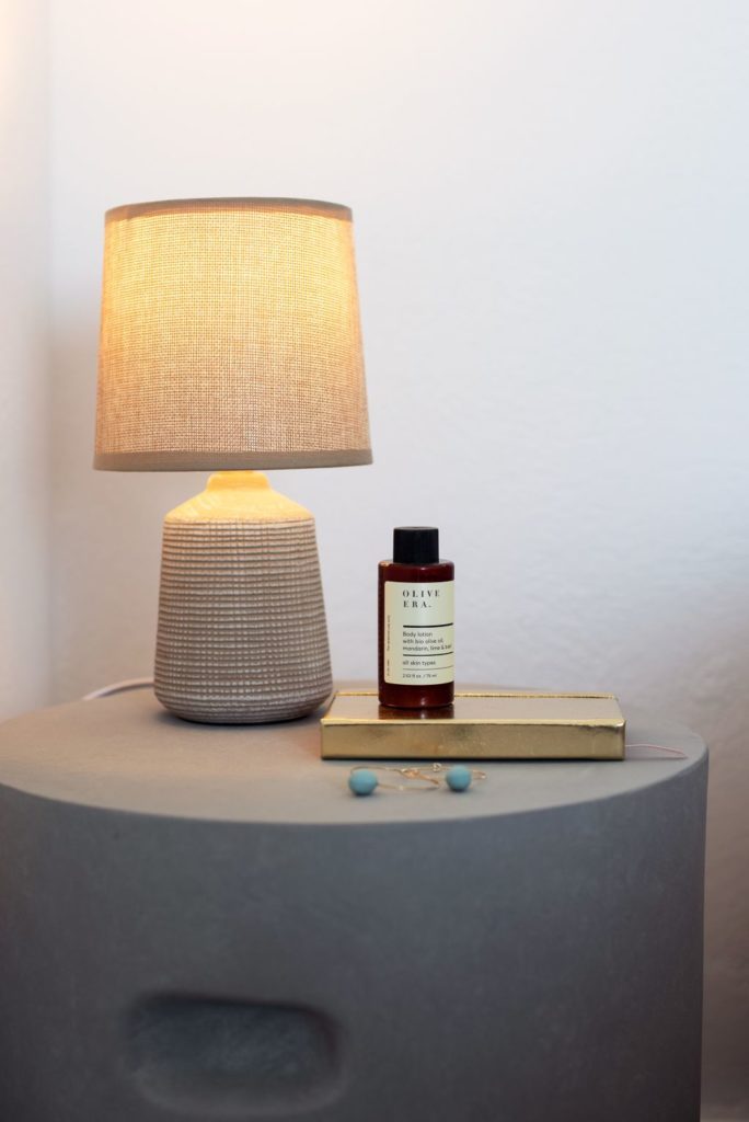 Bedside table with a linen-shaded lamp, a book and bathroom oil