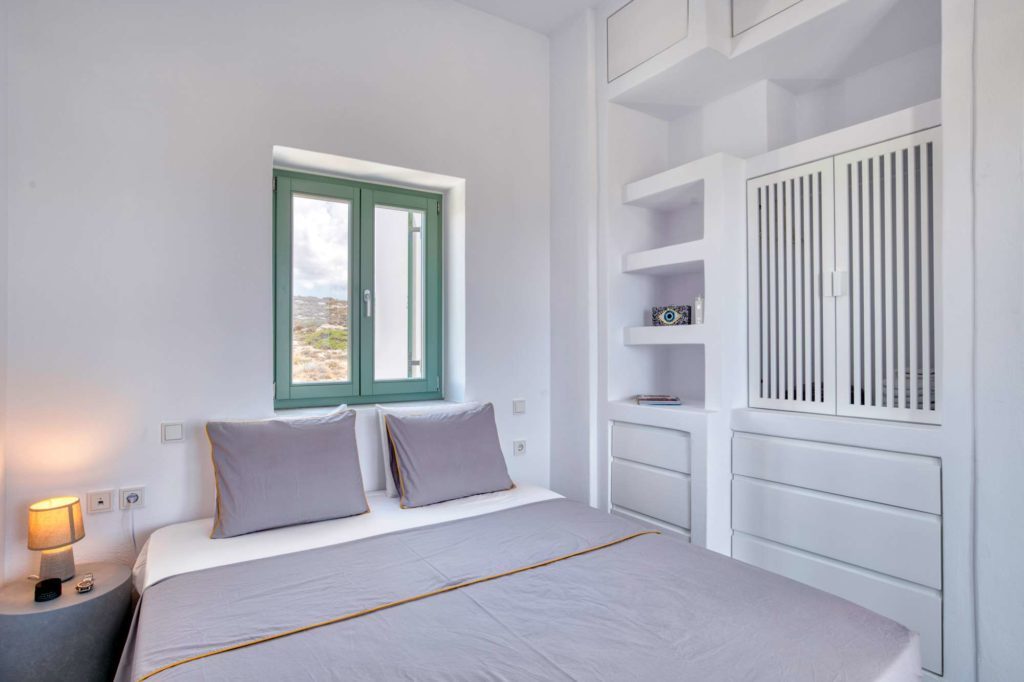 The queen bed with grey linen, built-in shelving and a green-framed window