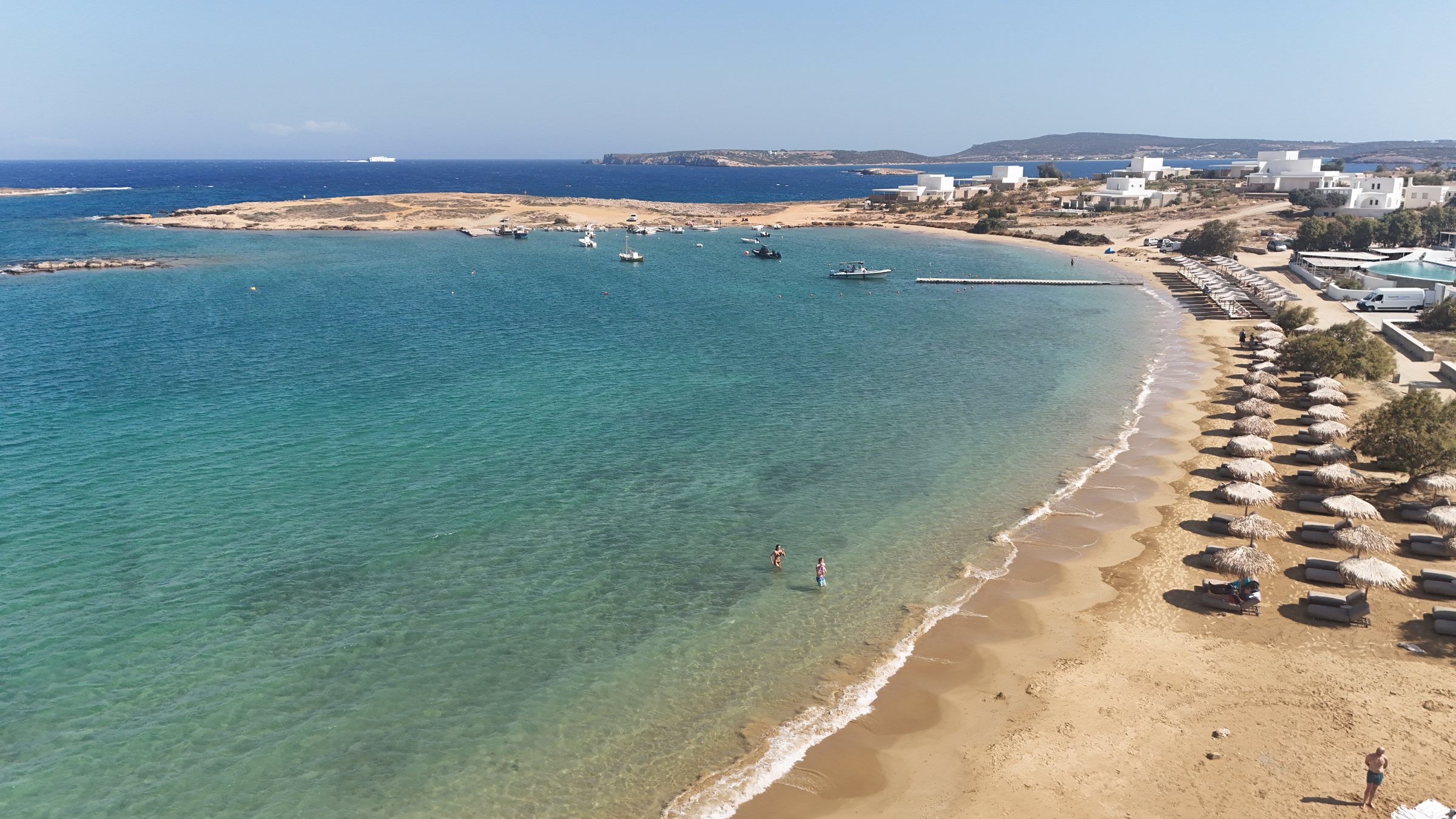 Agioi Anargyri from above: the sheltered turquoise bay, the sandy crescent lined with straw parasols, moored boats and the headland beyond